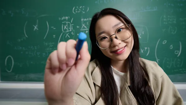 A student using a white marker pen to write a mathematical equation on a glass surface. They are wearing glass and a beige jacket, and they are stood in front of a blackboard which also features a mathematical equation.