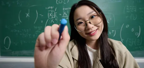 A student using a white marker pen to write a mathematical equation on a glass surface. They are wearing glass and a beige jacket, and they are stood in front of a blackboard which also features a mathematical equation.
