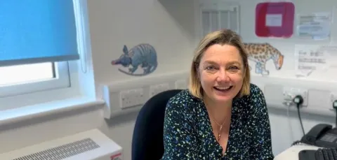 Professor Juliet Gray sitting at her desk, smiling. There are pangolin and leopard stickers on a white wall in the background. The window behind her is mostly covered by a light blue blind.