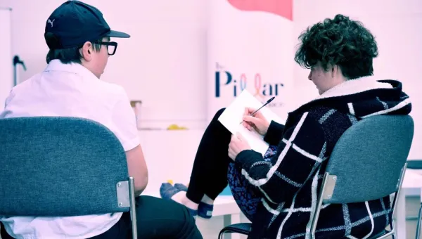 Two young adults sitting with their backs to the camera. One has their feet up on a table and is writing on a note pad resting on their knee. The other has a baseball cap on and is watching the person writing. there is a Pillar logo on the wall.