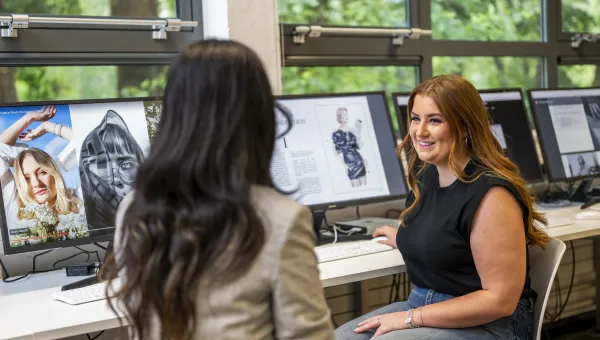 Two people are shown seated and in discussion, in front of wide screen computers.