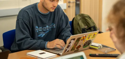 A student with short blonde hair sat at a desk with a laptop open. They are wearing a Reebok sweater, and several rings on their fingers.