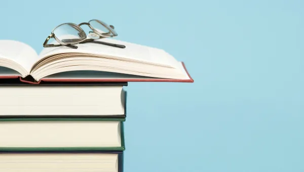 A stack of hardcover books with an open book on top and glasses resting on it against a blue background.