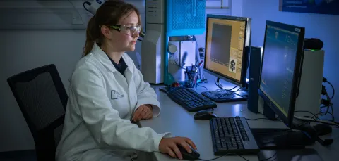 A Chemistry and Chemical Engineering technician in a lab, wearing a lab coat and goggles while sat at a desk using a computer.