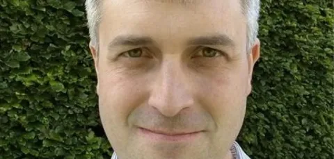 A man with short gray hair wearing a striped shirt, standing in front of a green hedge and looking at the camera.