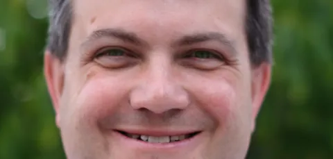 A close-up of a man with short dark hair, wearing a light blue collared shirt. They are smiling. They are stood outdoors, with blurred green trees in the background.