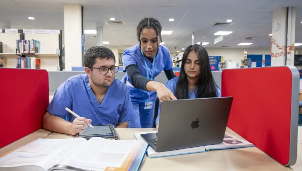 Medicine students wearing blue scrubs, study together in a library, with a laptop, tablet and reference book