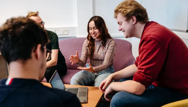 Four people sitting around a small table in a casual meeting setting. One person is speaking and gesturing with hands, while others are listening attentively. A laptop is open on the table, and the group is seated on cushioned chairs in a bright room with a whiteboard in the background