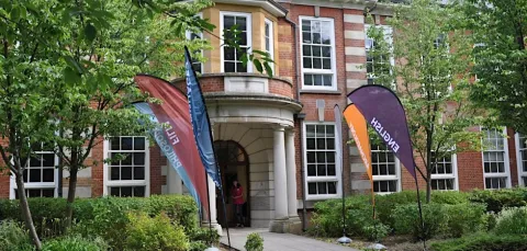 A brick building with large white-framed windows and a columned entrance is surrounded by greenery. Colorful flags labeled “English” and “Film” stand along the pathway leading to the doorway.