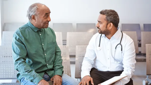 A doctor consulting a patient in a hospital waiting room. The doctor is holding a clipboard and has a stethoscope around their neck.