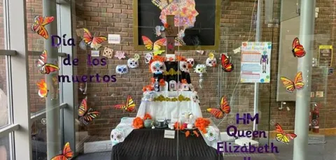 A Día de los Muertos altar is displayed behind glass, decorated with colorful paper butterflies, sugar skulls, flowers, and candles. Text on the glass reads “Día de los muertos” and “HM Queen Elizabeth II.”