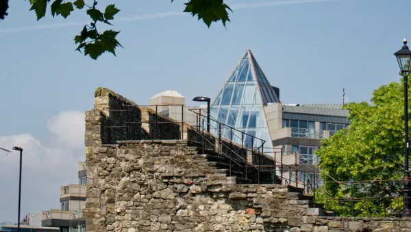 View of a historic stone wall with staircase and metal railings in the foreground, juxtaposed against a modern building featuring a glass pyramid roof. The scene includes leafy tree branches above and a traditional street lamp on the right, highlighting the contrast between medieval and contemporary architecture in an urban setting.
