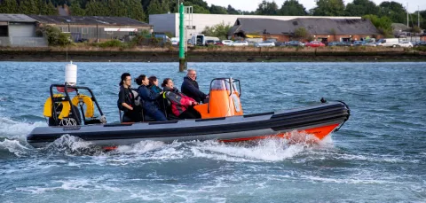 Students wearing life jackets ride a small orange RIB boat across the water during a summer school activity.