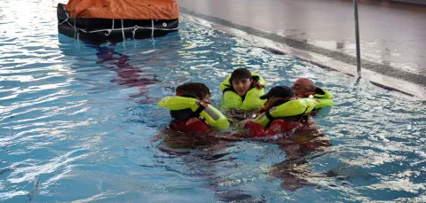 Four people wearing bright yellow life jackets and red immersion suits are floating in a swimming pool, huddled together in a survival position. 