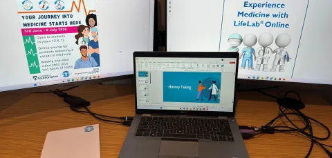 A desk setup with two large monitors and an open laptop. The monitors display information about the Experience Medicine Online course.