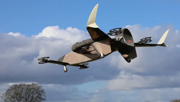 A drone with vertical stabilisers and multiple rotors flying through a blue sky with scattered clouds, above a green field dotted with trees.