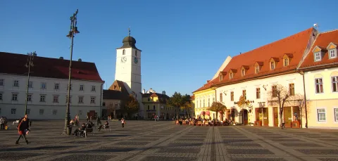 Small, Romanian market square with a clock tower, cobbled paving and painted white buildings, blue sky overhead.