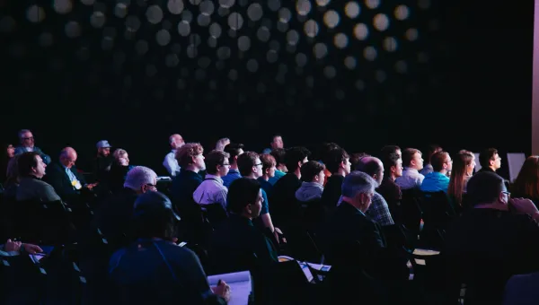 A three quarter view from behind of conference attendees sat in rows. The room is mostly dark and there is light coming from the front of the room, out of the shot. 
