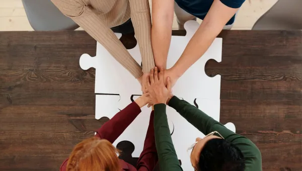 Hands of four people stacked over a white puzzle piece on a wooden table.