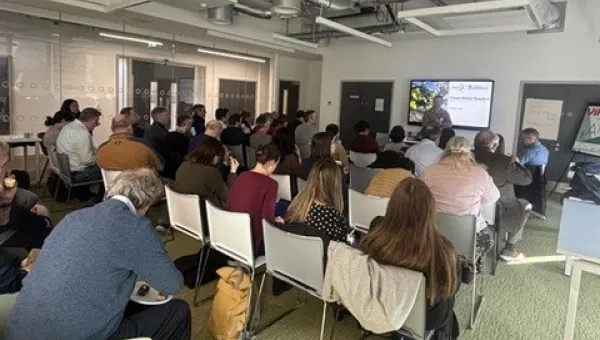 A meeting room with attendees seated in rows facing a speaker and a presentation screen.