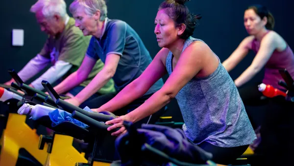 People taking part in an indoor cycling class, seated on stationary bikes in a dimly lit studio with colourful lighting.