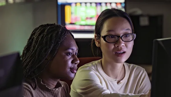 Two students seated together in the Bloomberg suite. One student gestures towards something on a computer screen.