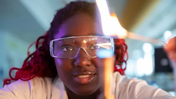 A chemistry student wearing lab coat and safety glasses focuses intently on the Bunsen burner she's igniting, looking determined and content