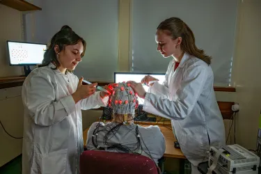 Two students adjust electrodes on a brain cap with red lights in a lab.