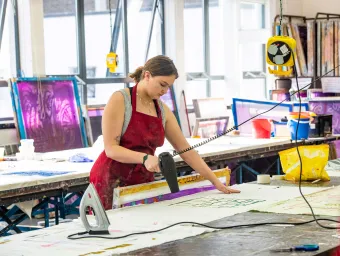 A student uses a hairdryer to dry a large piece of artwork in an art studio.