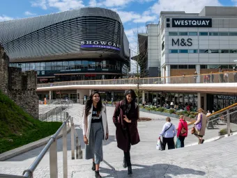 Two students talk whilst walking across a large paved area with several large shops.