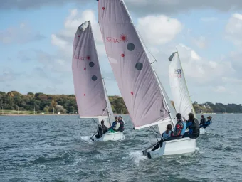 Three small sailing boats, each with a crew of three, on open water beneath blue skies. An area of trees is visible in the background.
