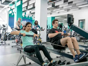 Group of students working out on rowing and cycling machines in a bright, modern gym.