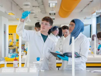 Two students wearing lab coats and protective glasses conduct an experiment. One holds up a glass beaker, while the other takes notes. They are in a laboratory, surrounded by a variety of chemistry equipment.