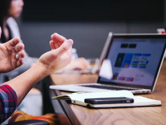 Laptop on a table with notepad and phone, user's hands in foreground 