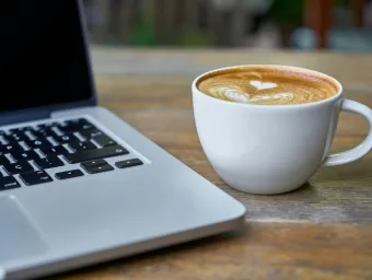 Stock image of a laptop open and a cup of coffee on a table. Image taken by pixabay.
