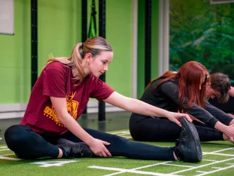  Students sitting on the floor in the gym with one leg out in front of them. They are reaching to hold on to their foot and stretching their leg muscle. 