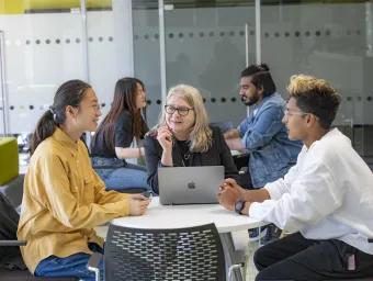 Two students and an advisor talking around a table. The advisor has a laptop open on the desk. There are other people sitting around tables in the background.