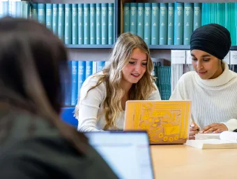 Two students discussing work in a library with a yellow laptop.