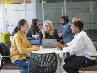 A group of three people sat around a table. They're talking in the group with a share laptop in the middle.