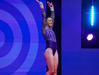 Elite athlete Emily Burke in a purple and silver leotard stands with her arms raised, smiling beneath stage lights after completing her routine.
