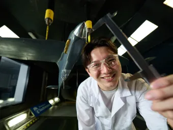 A smiling University of Southampton student in a lab coat and safety glasses, holding a caliper towards the camera. Behind them is a 1/6th scale Spitfire model that was put together for a 3rd year individual project in the University's R J Mitchell Wind Tunnel on Highfield Campus.