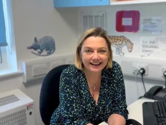 Professor Juliet Gray sitting at her desk, smiling. There are pangolin and leopard stickers on a white wall in the background. The window behind her is mostly covered by a light blue blind.
