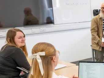 A teacher sits on a desk holding papers while three students face them, one using a laptop. A whiteboard behind the teacher displays the phrase "POINT OF VIEW."