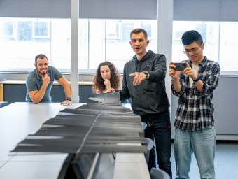 In a classroom setting, a staff member demonstrates wave propagation using a specialized device, while three researchers observe attentively.