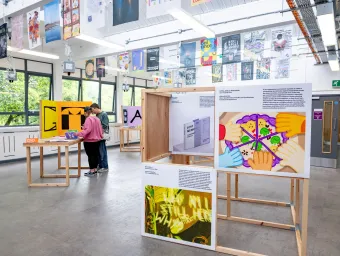 An exhibition space with large windows displays colorful graphic design projects mounted on wooden frames, featuring bold illustrations and text. Additional posters hang from the ceiling, and tables with more design work are arranged throughout the room.