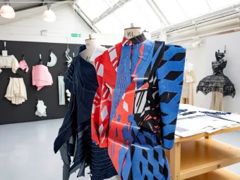 Two dress forms display bold, patterned garments, one in blue and red abstract shapes and the other in dark blue layered fabric. In the background, a black wall holds various fabric samples and sculptural pieces, with another black structured dress visible to the right.