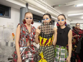 Three models stand in a brightly lit backstage area wearing bold, patterned outfits made from mixed fabrics including plaid and vibrant prints. The background shows clothing racks and other people preparing for what appears to be a fashion event.