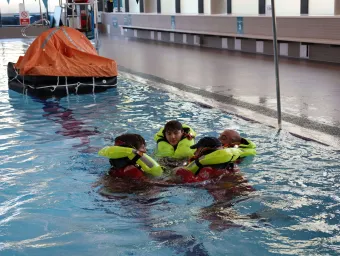 Four people wearing bright yellow life jackets and red immersion suits are floating in a swimming pool, huddled together in a survival position. 