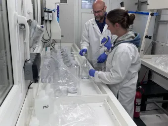 Two people in a laboratory preparing water samples, wearing lab coats and gloves, and handling bottles and equipment on a long workbench.