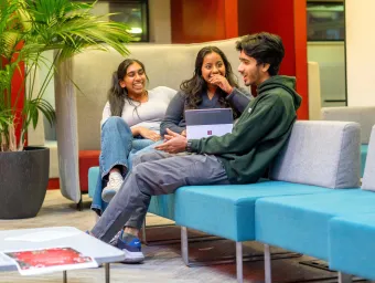 Three students sat of a sofa in student halls with an ipad. They're talking and smiling.